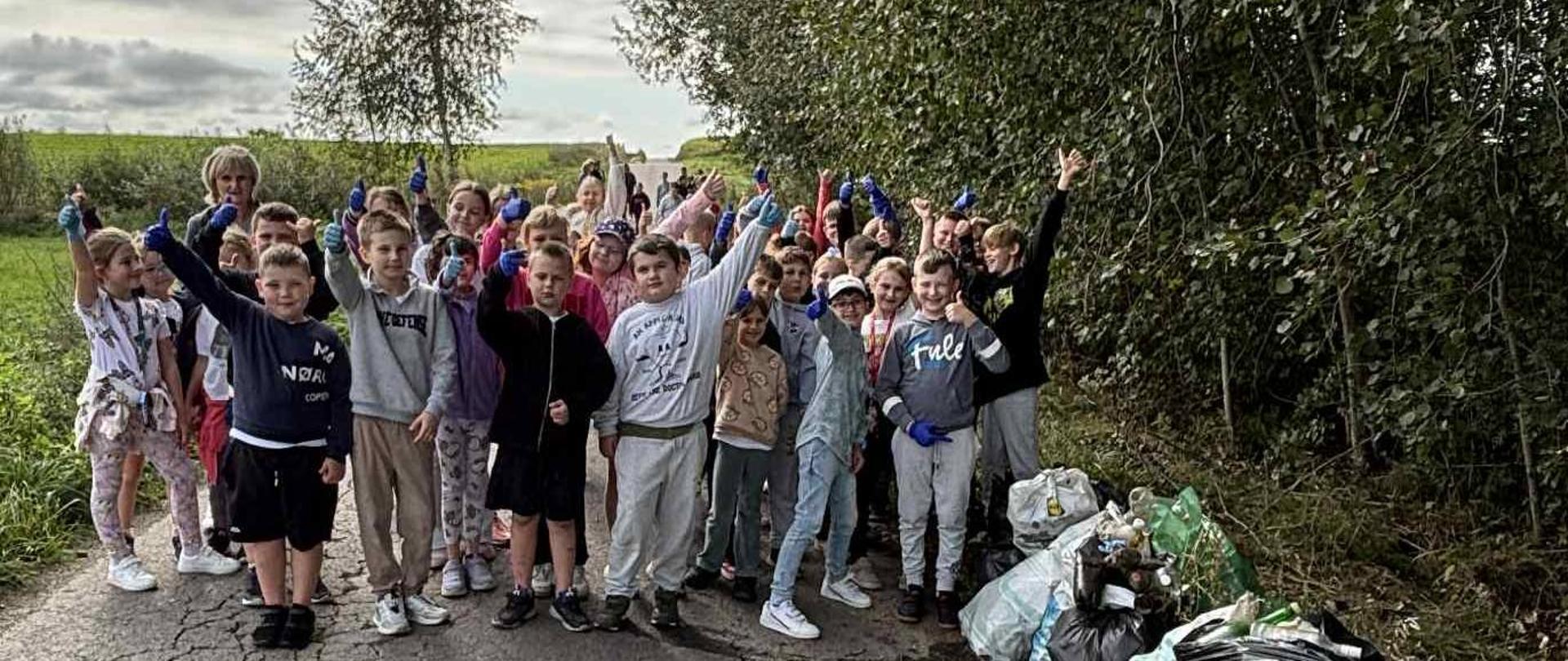 Grupa dzieci pozuje do wspólnego zdjęcia. Wszyscy znajdują się na drodze, obok leżą worki z śmieciami. Dzieci uśmiechają się i machają rękoma.