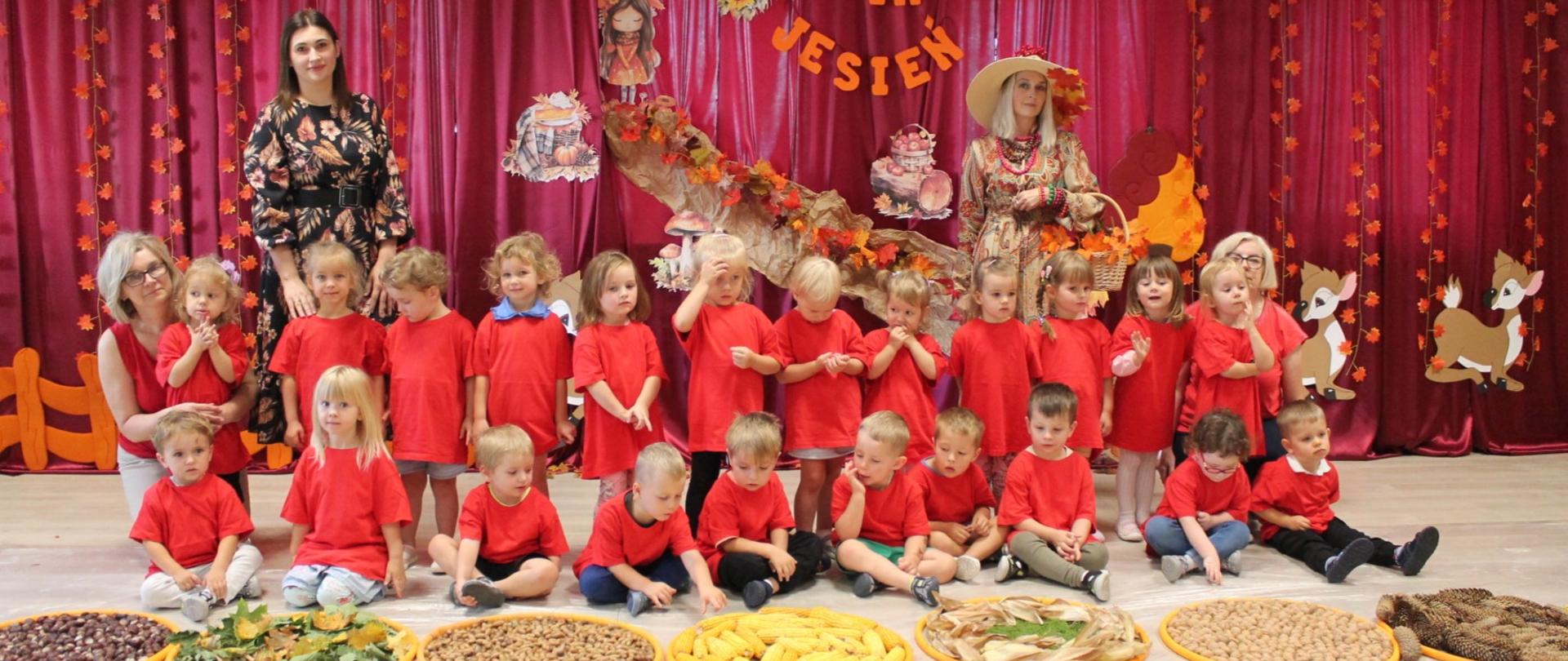 3 kobiety i grupa dzieci w czerwonych koszulkach na tle napisu WITAJ JESIEŃ 