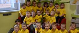 Dzieci z grupy Delfinki ubrane w żółte koszulki, siedzą w trzech rzedach i trzymają bukiet kwiatów