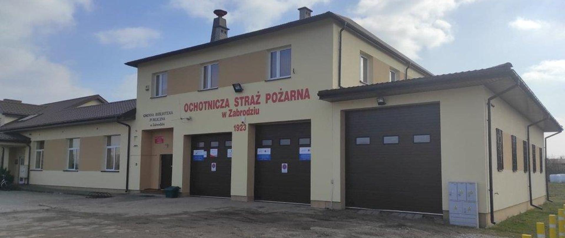 Piętrowy budynek w kolorze żółtym z czerwonymi napisem nad parterem Ochotnicza Straż Pożarna w Zabrodziu z trzema dużymi brązowymi bramami garażowymi.