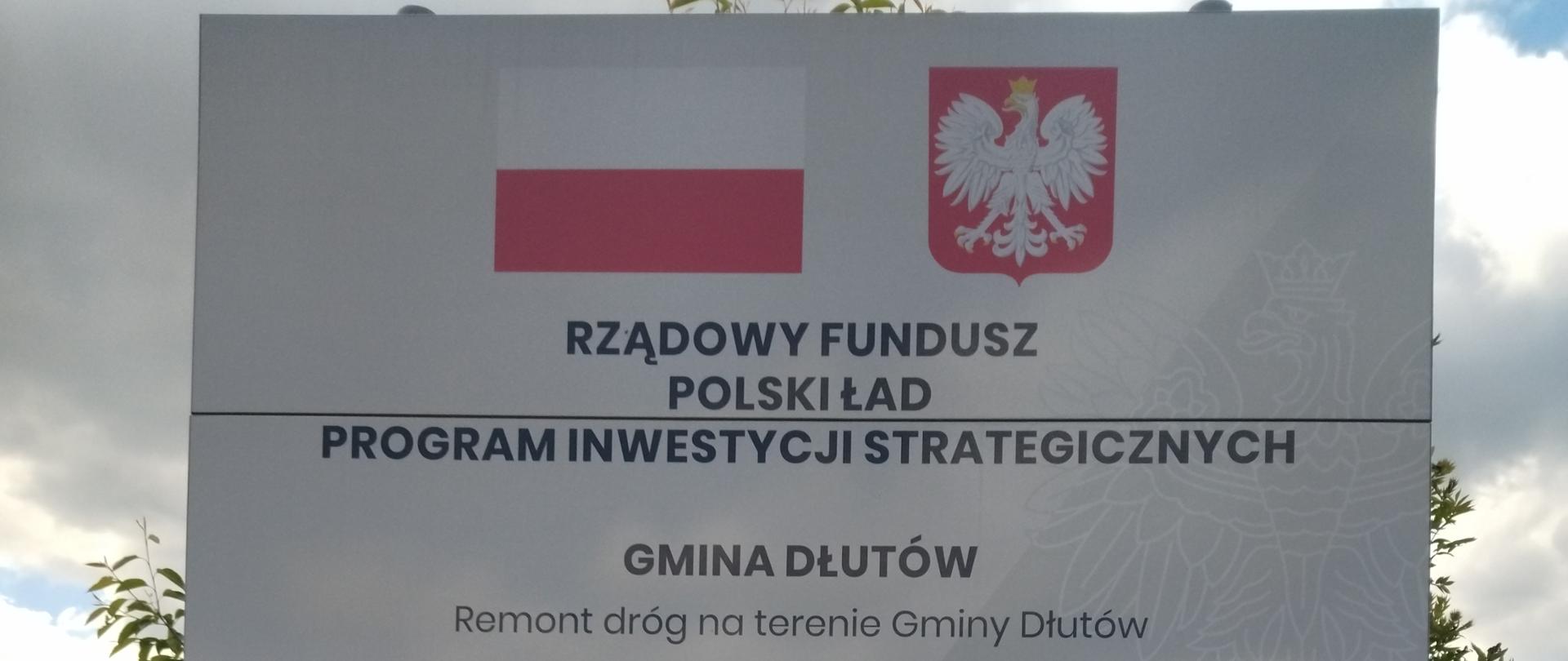 Tablica informacyjna na tle pochmurnego nieba, promująca inwestycję w Gminie Dłutów. Widnieją na niej polskie godło i flaga oraz napisy: "Rządowy Fundusz Polski Ład Program Inwestycji Strategicznych" i "Gmina Dłutów Remont dróg na terenie Gminy Dłutów". Podana kwota dofinansowania to "3.476.035,65 zł".