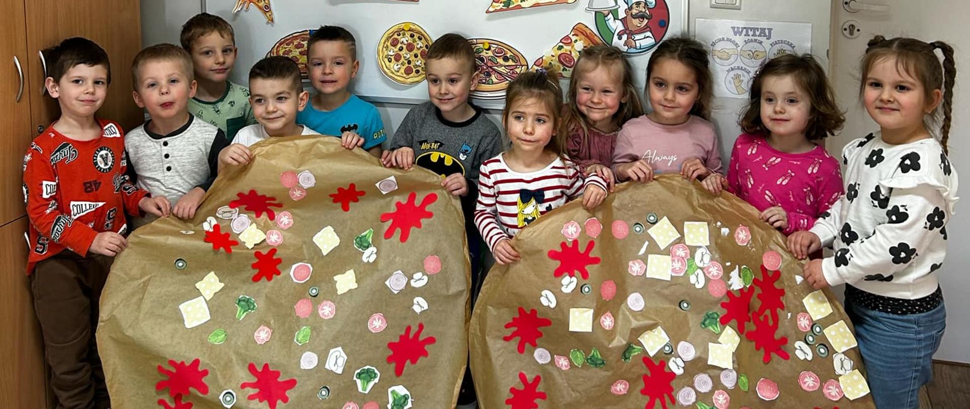 Grupa dzieci w przedszkolu, które stoją w rzędzie i trzymają duże, wykonane z papieru „pizze”. 
