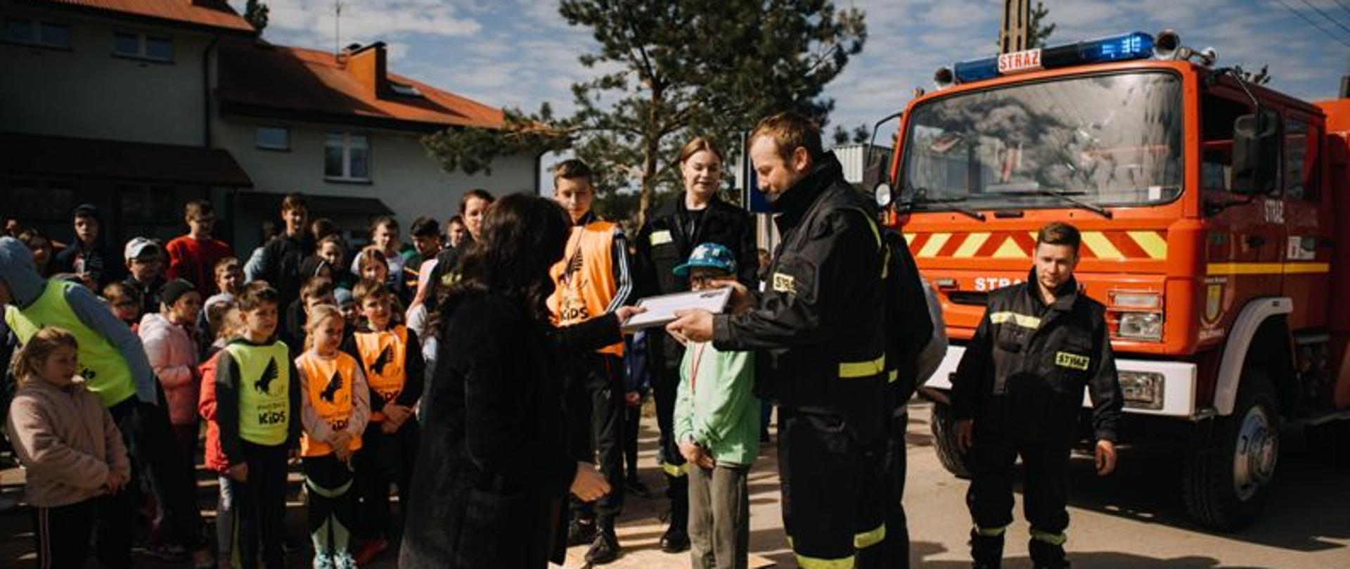 Zdjęcie przedstawia na pierwszym planie Panią Dyrektor, która wręcza dla strażaka dyplom. Podium ustawione przed samochodem strażackim koloru czerwonego na którym stoją uczniowie i strażacy. Uczniowie całej szkoły stoją z boku i obserwują wręczanie dyplomów. 