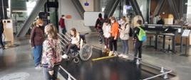 Dziewczynka na wózku inwalidzkim jeździ po czarnej podłodze. Wokół inne dzieci