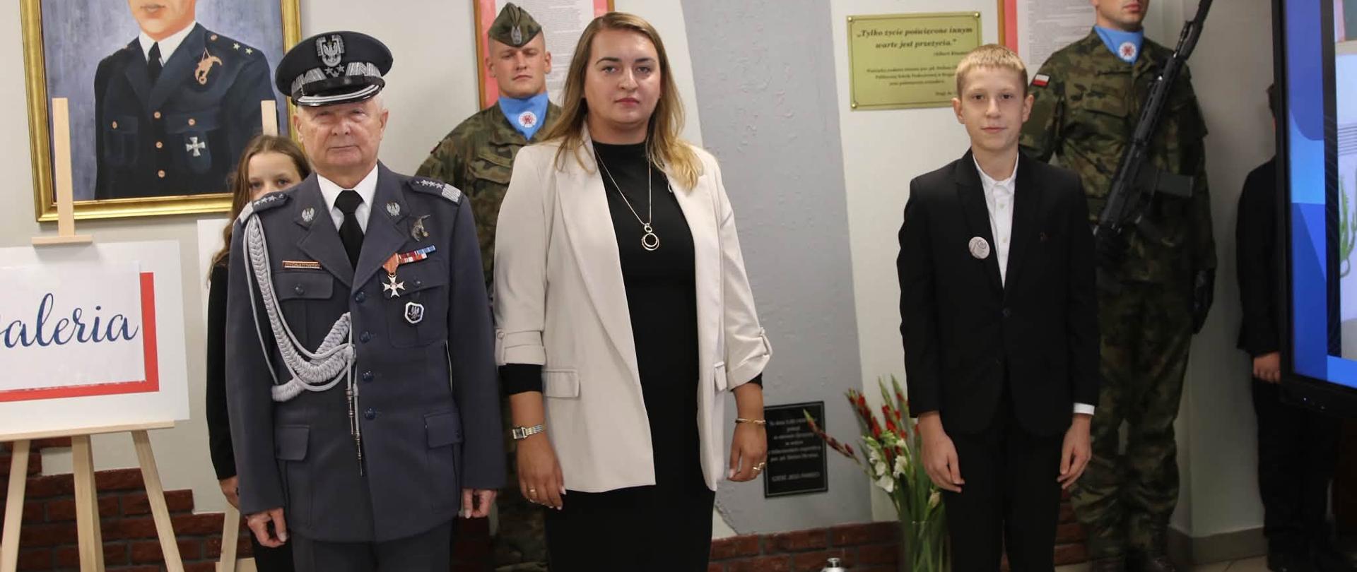 Uroczystość w sali, podczas której odbywa się oficjalna część wydarzenia. Na zdjęciu widoczni są przedstawiciele służb mundurowych, uczestnicy uroczystości oraz młodzież szkolna. W tle znajduje się wystawa poświęcona historii i tradycjom służb mundurowych.