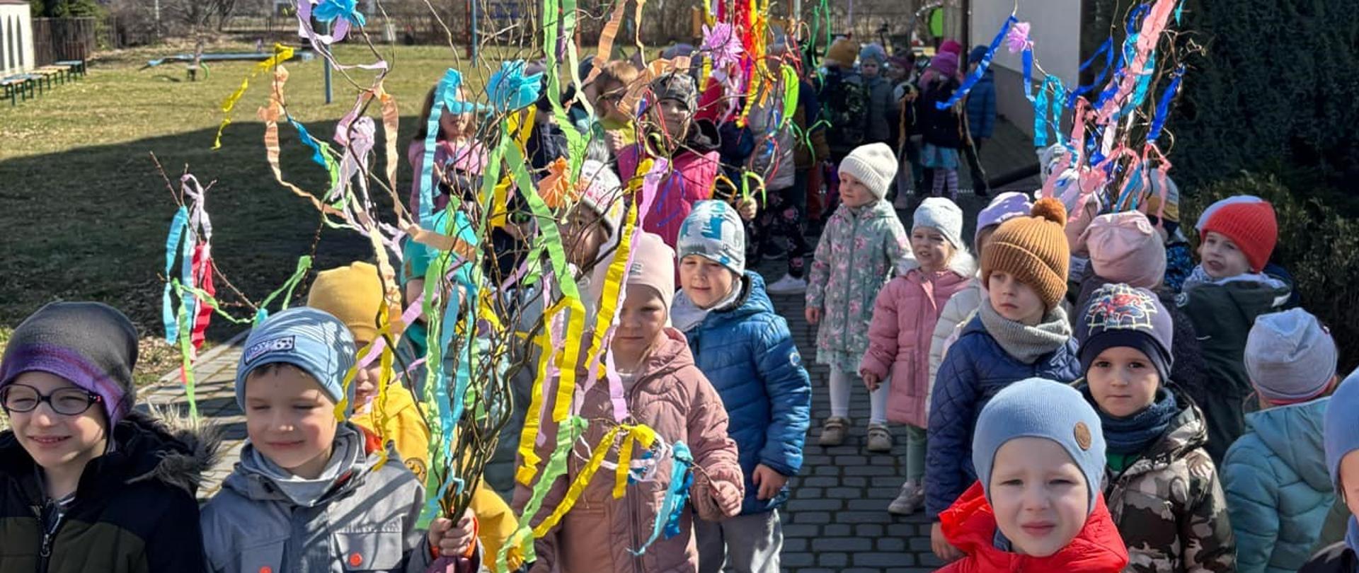 Przedszkolaki w barwnym korowodzie z gaikami spacerują w okolicy przedszkola witając pierwszy dzień Wiosny.
