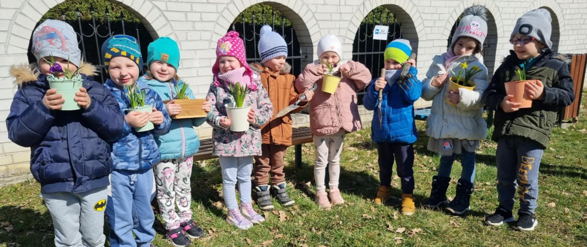 Grupa dzieci stoi na przedszkolnym podwórku. Dzieci trzymają kolorowe doniczki z kwiatami, które właśnie same zasadziły, a także narzędzia ogrodnicze. 