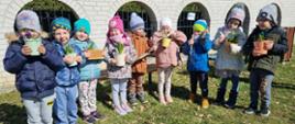 Grupa dzieci stoi na przedszkolnym podwórku. Dzieci trzymają kolorowe doniczki z kwiatami, które właśnie same zasadziły, a także narzędzia ogrodnicze. 
