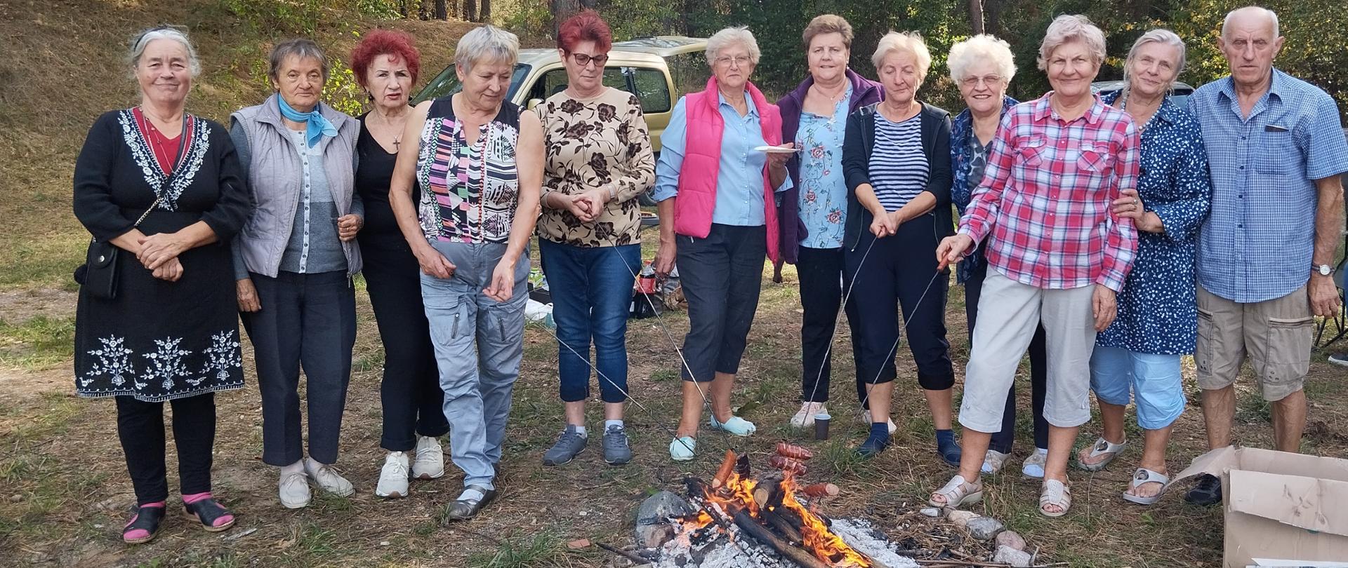 członkowie klubu senior+, piekący kiełbaski nad ogniskiem oraz pozujący do wspólnego zdjęcia