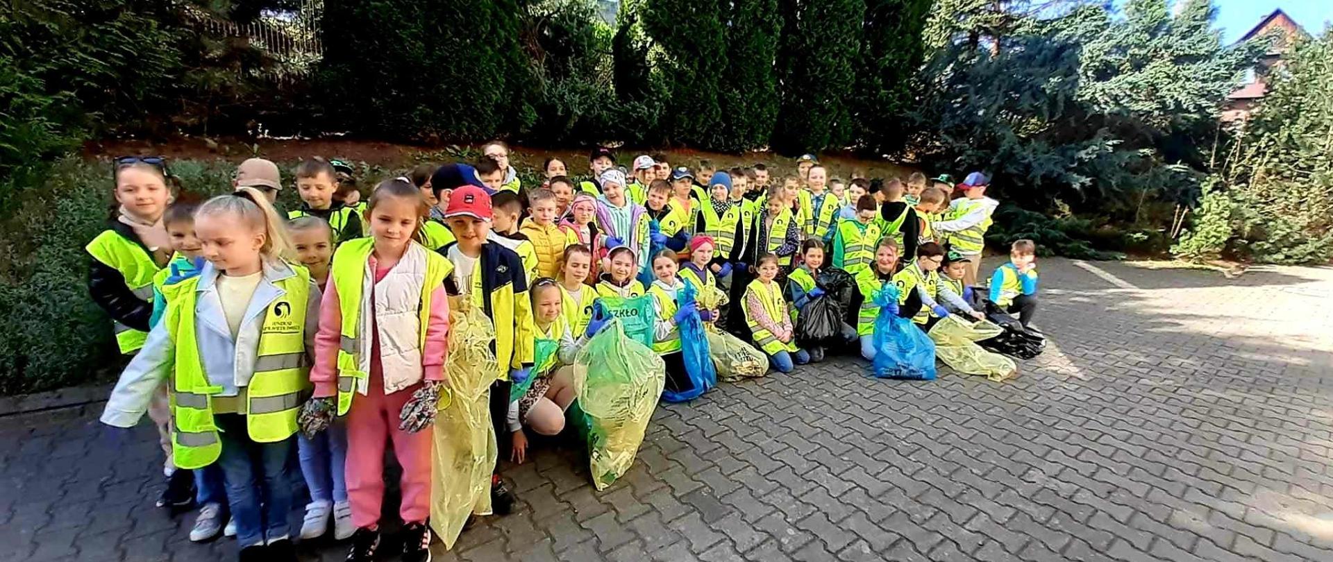 Uczniowie klas I- III ustawieni przed budynkiem hali sportowej, ubrani w odblaskowe kamizelki oraz jednorazowe rękawiczki uczestniczą w akcji sprzątania śmieci.