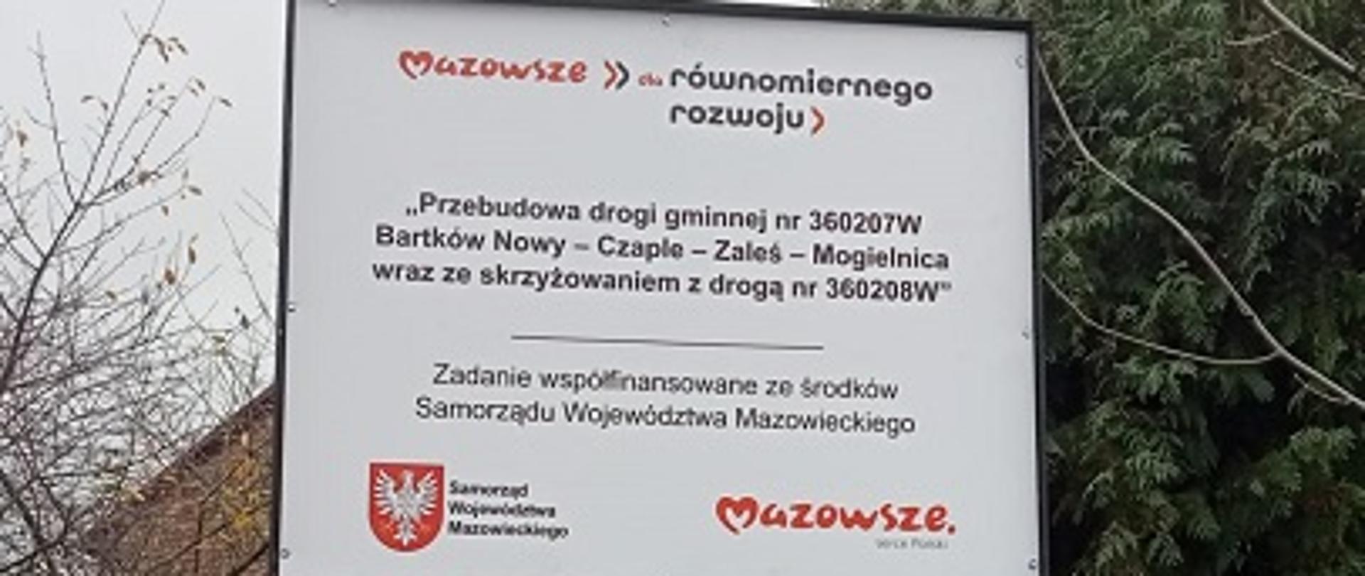 tablica informacyjna o projekcie, w tle kapliczka przydrożna, studnia i dom z czerwonej cegły