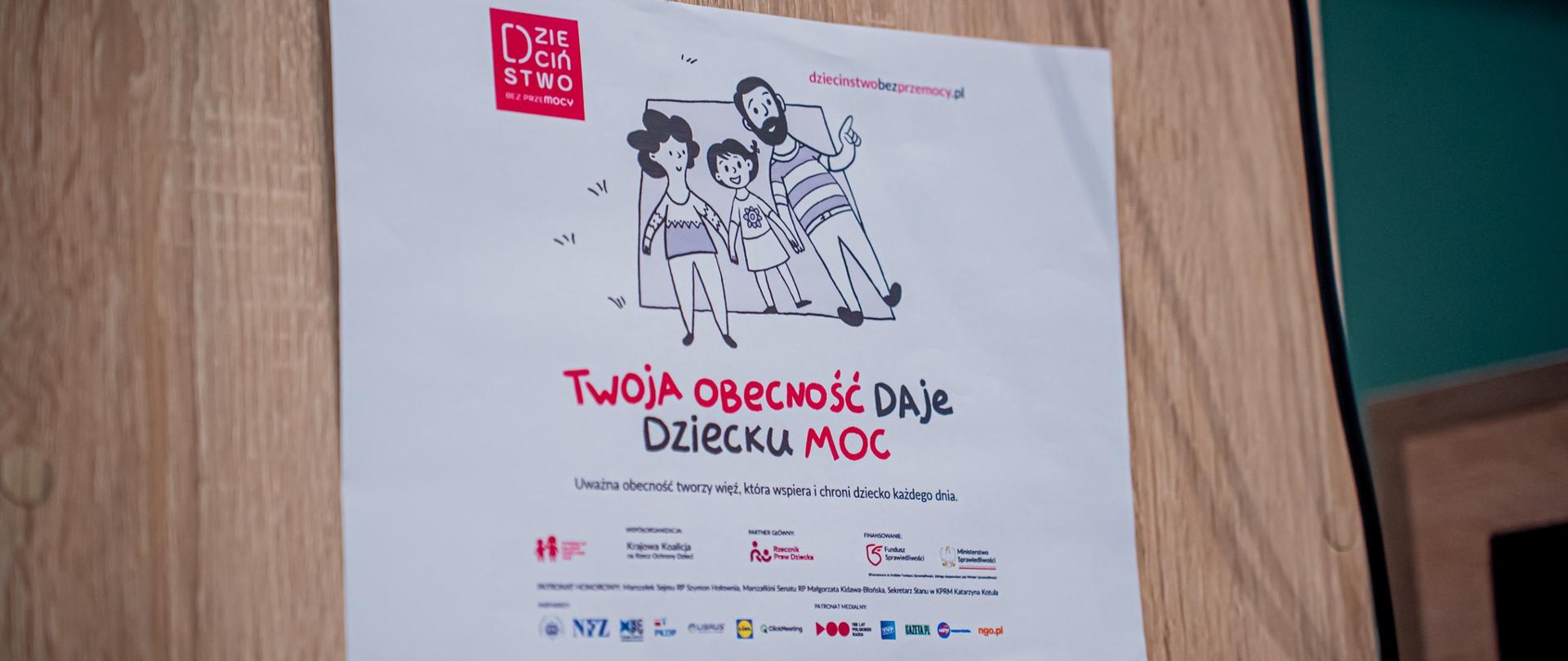 Zdjęcie przedstawia plakat kampanii społecznej „Dzieciństwo bez przemocy” z hasłem „Twoja obecność daje dziecku moc”. Grafika ukazuje rodzinę – rodziców i dziecko – symbolizującą bliskość, troskę i bezpieczeństwo. Plakat przypomina, że uważna obecność dorosłych buduje więź, która wspiera i chroni dziecko każdego dnia.