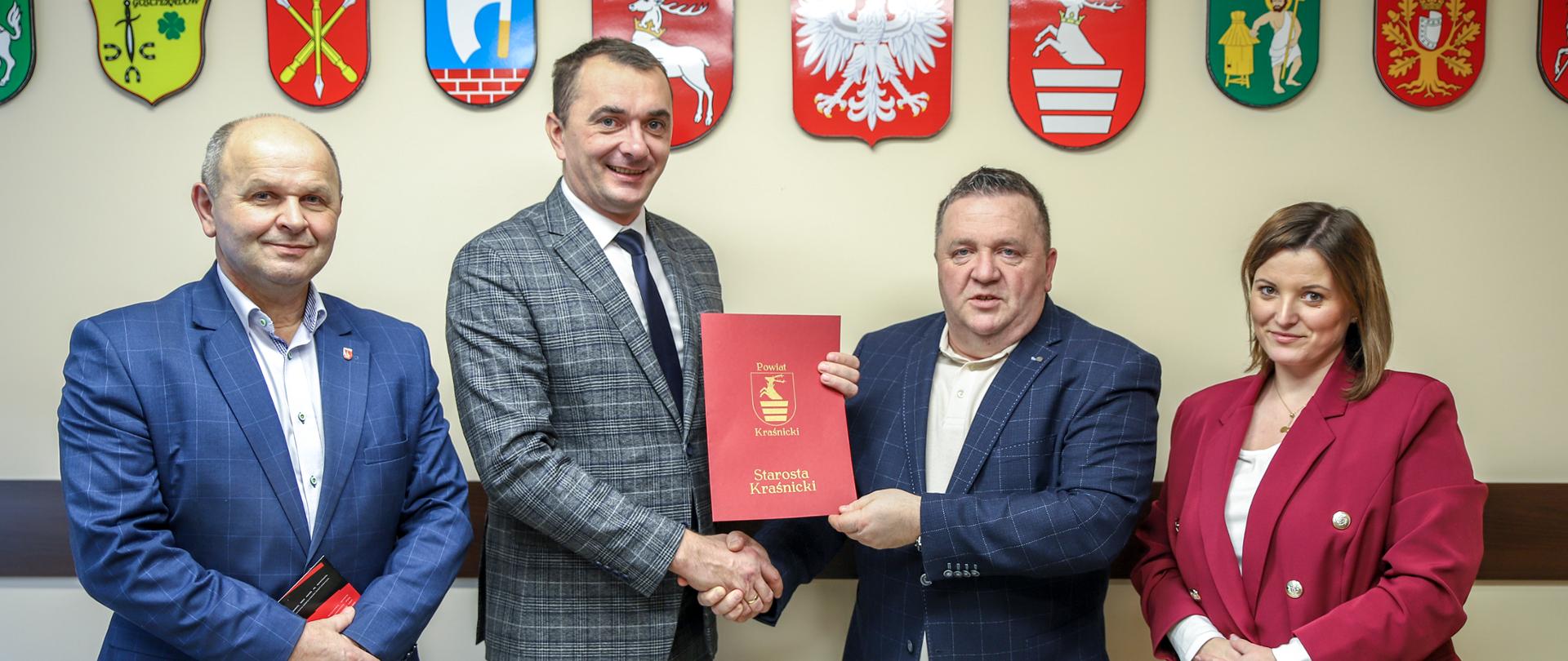 Pamiątkowe zdjęcie z podpisania umowy uwieczniające moment przekazania podpisanych dokumentów. W tle znajdują się godło oraz herby województwa lubelskiego, powiatu kraśnickiego i gmin z jego terenu.