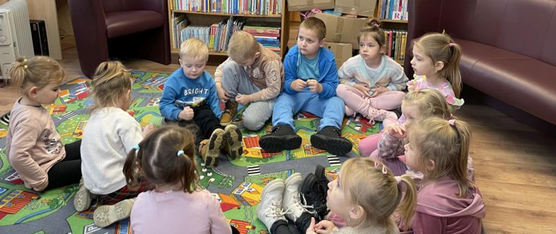 Grupa dzieci siedzi na dywanie, w tle półki z książkami.