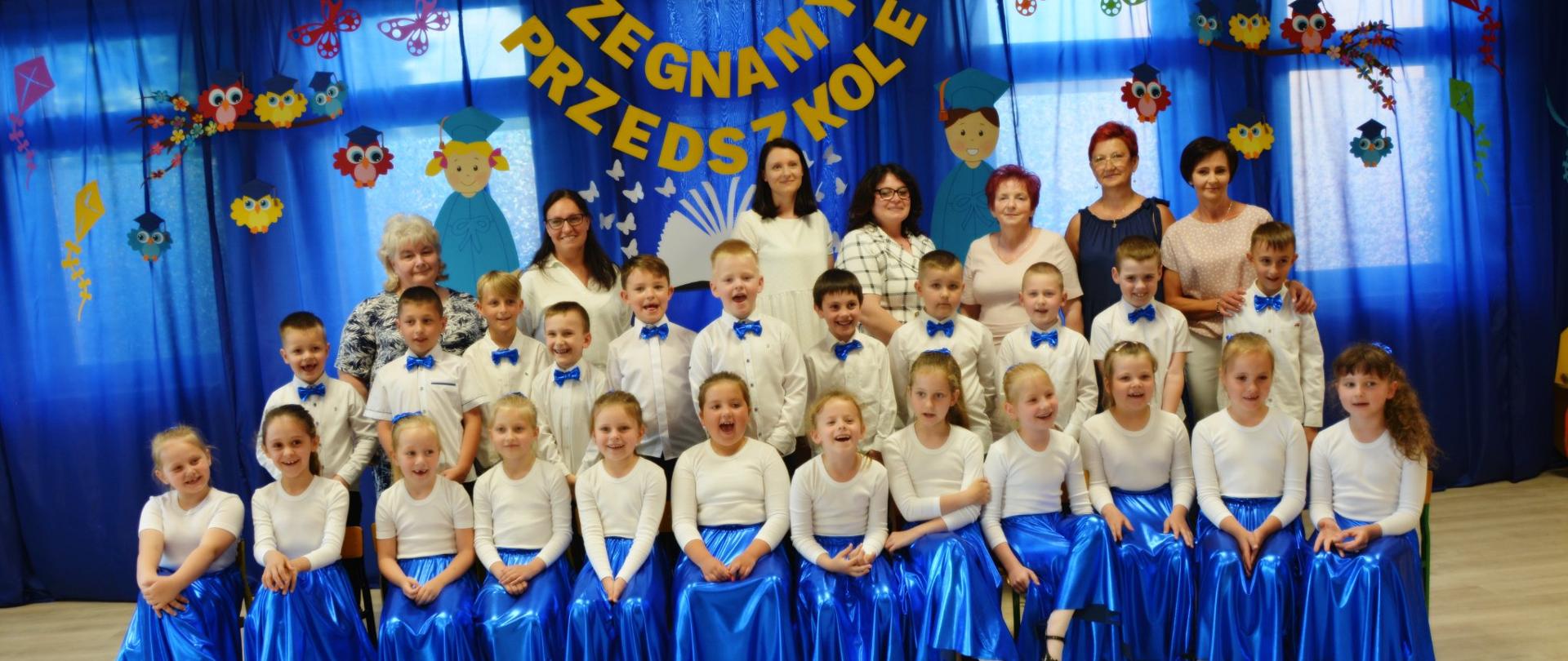 Dzieci w strojach galowych na tle napisu żegnamy przedszkole