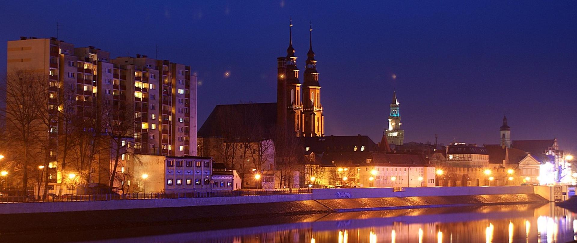 Nocna panorama Opola uchwycona z perspektywy Odry. Centralnym punktem fotografii jest podświetlona katedra Świętego Krzyża. W tle po prawej stronie wieża ratusza.