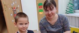 Chłopczyk i jego mama siedzą przy stole, wspólnie tworząc świąteczne ozdoby - papierową torbę z bożonarodzeniowym akcentem