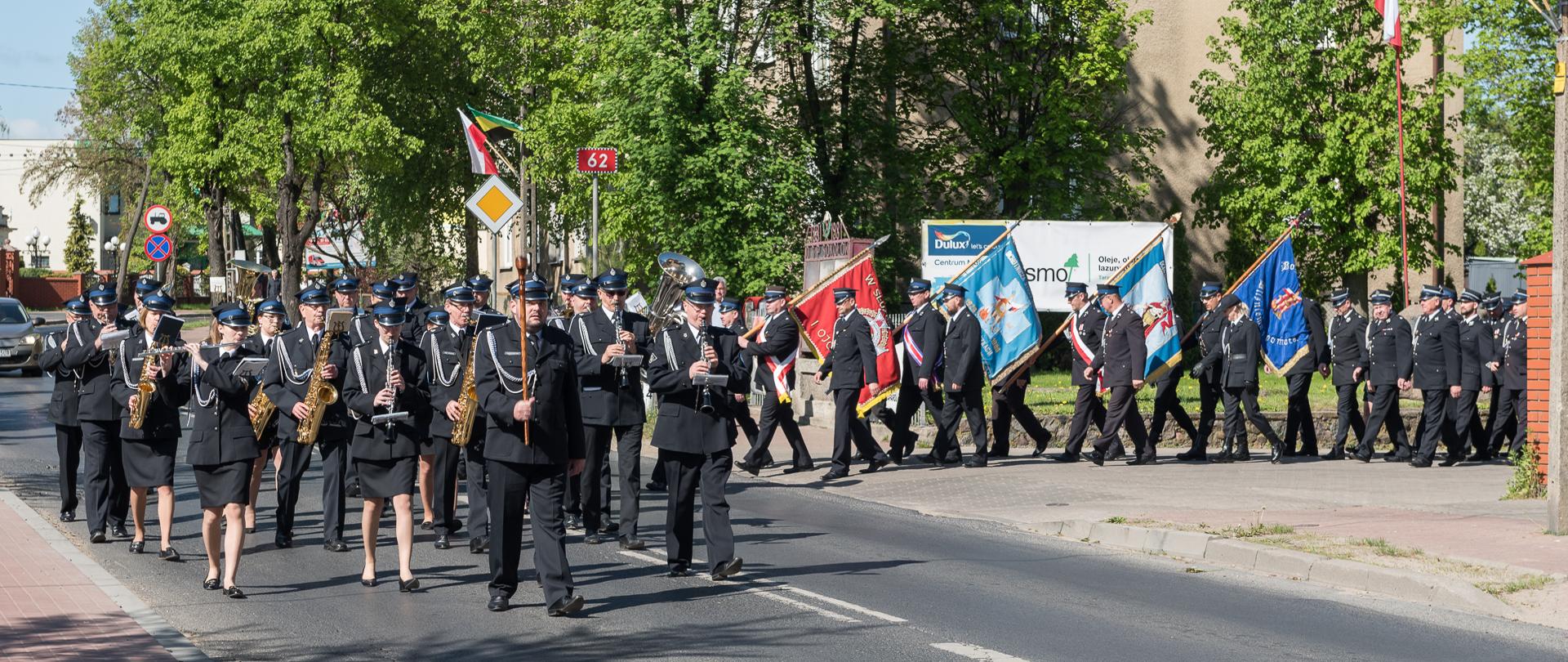 Na fotografii widać kolumnę zmierzającą ulicą miasta Sokołów Podlaski na uroczystą Mszę Świętą. Kolumnę prowadzi orkiestra dęta, a za nią idą poczty sztandarowe oraz strażacy z jednostek OSP w mundurach galowych.