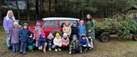 Dzieci i nauczycielki pozują do wspólnego zdjęcia przy czerwonym samochodzie udekorowanym lampionami, obok wóz z choinkami.
