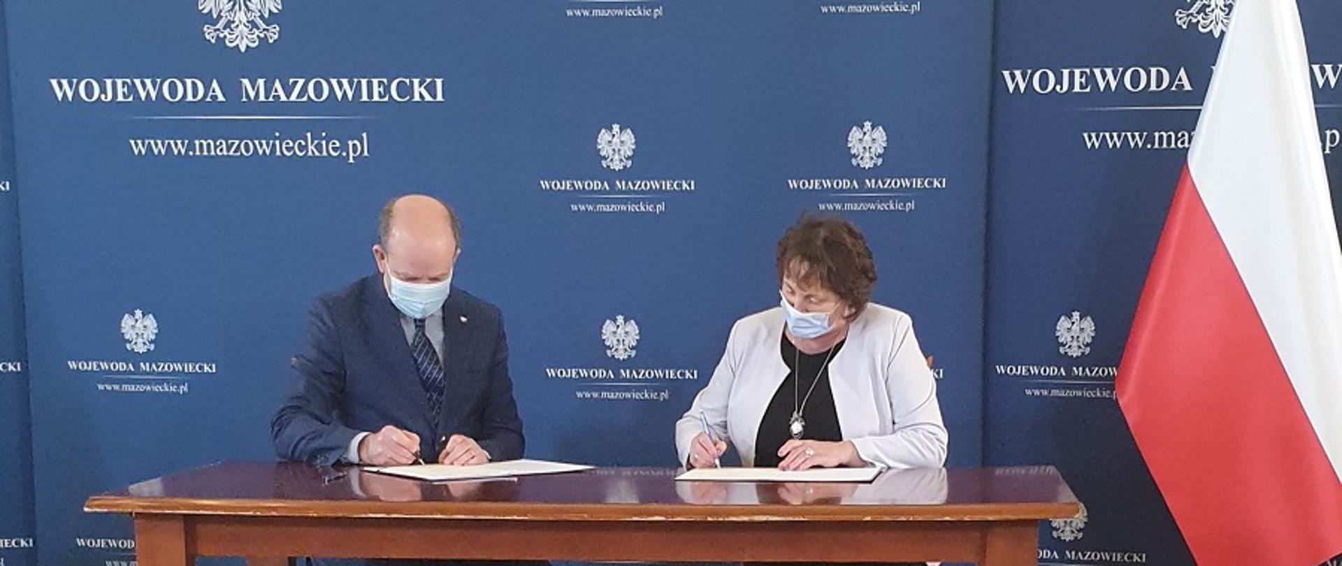 Starosta Elżbieta Sadowska podpisuje z wojewodą mazowieckim Konstantym Radziwiłłem umowę na dofinansowanie do przewozów autobusowych o charakterze użyteczności publicznej. W tle niebieska ścianka z logo wojewody oraz polska flaga. Podpisujący siedzą przy drewnianym stole