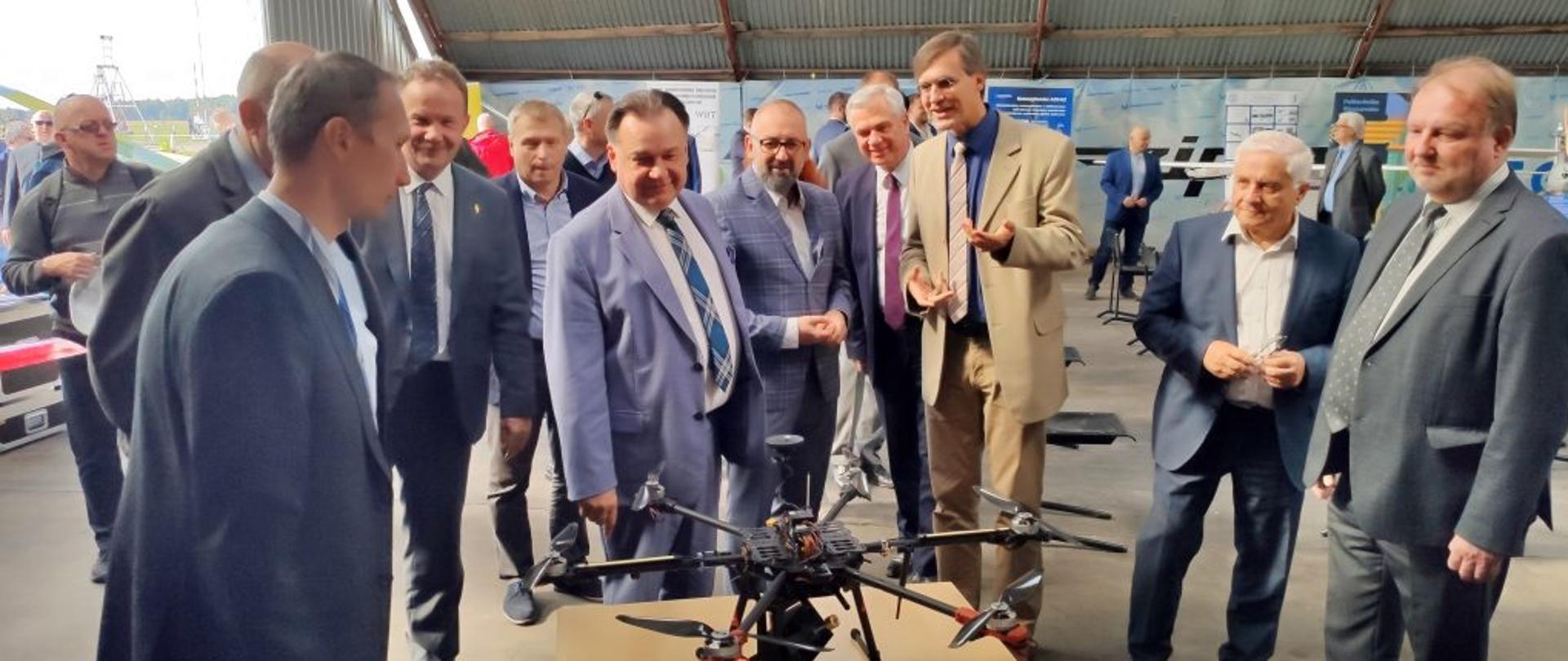 Zdjęcie przedstawia uczestników uroczystości oraz pracowników Politechniki Warszawskiej prezentujących gościom możliwości technologiczne latających urządzeń bezzałogowych