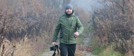 Mężczyzna w zielonej kurtce, czapce i kaloszach idzie ścieżką wśród jesienno-zimowej roślinności, trzymając w ręce aparat fotograficzny z teleobiektywem.