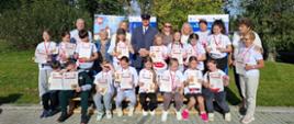 Młodzi uczestnicy imprezy wraz z organizatorami i zaproszonymi gośćmi na tle banerów promocyjnych, ubrani w białe koszulki okolicznościowe. Osoby trzymają w rękach dyplomy, a na szyjach mają zawieszone medale.