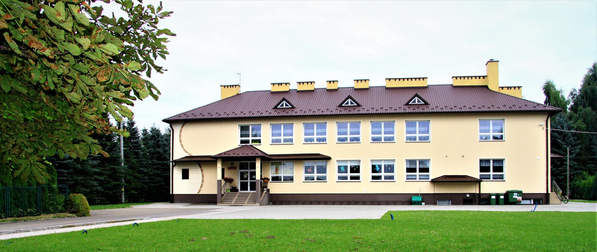 Widoczny front szkoły wraz z wejściem głównym widoczny trawnik i parking przed szkołą elewacja w jasnym kolorze okna białe dach brązowy w sąsiedztwie szkoły widoczne zadrzewienia po prawej i po lewej