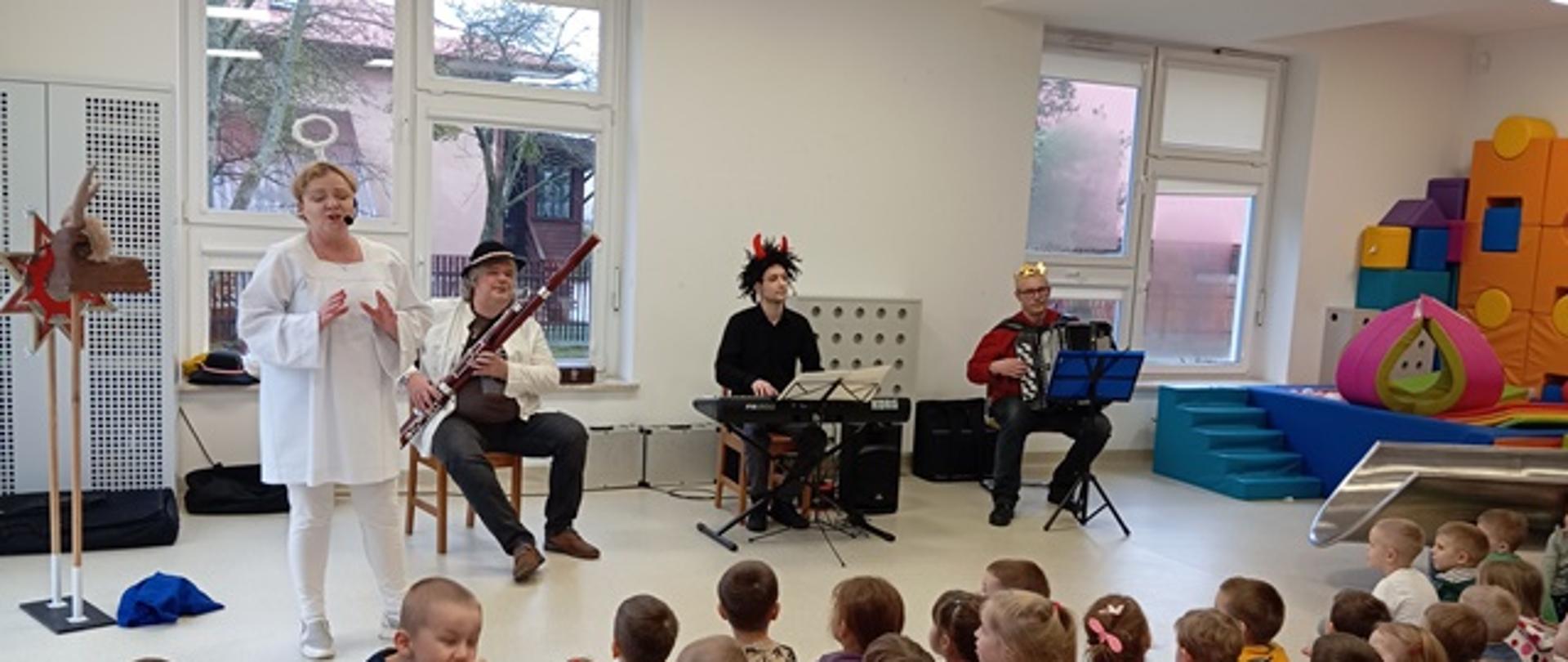 Zdjęcie prezentuje dzieci biorące udział w audycji muzycznej oraz teatrzyku. Przed grupą siedzących dzieci występują aktorzy