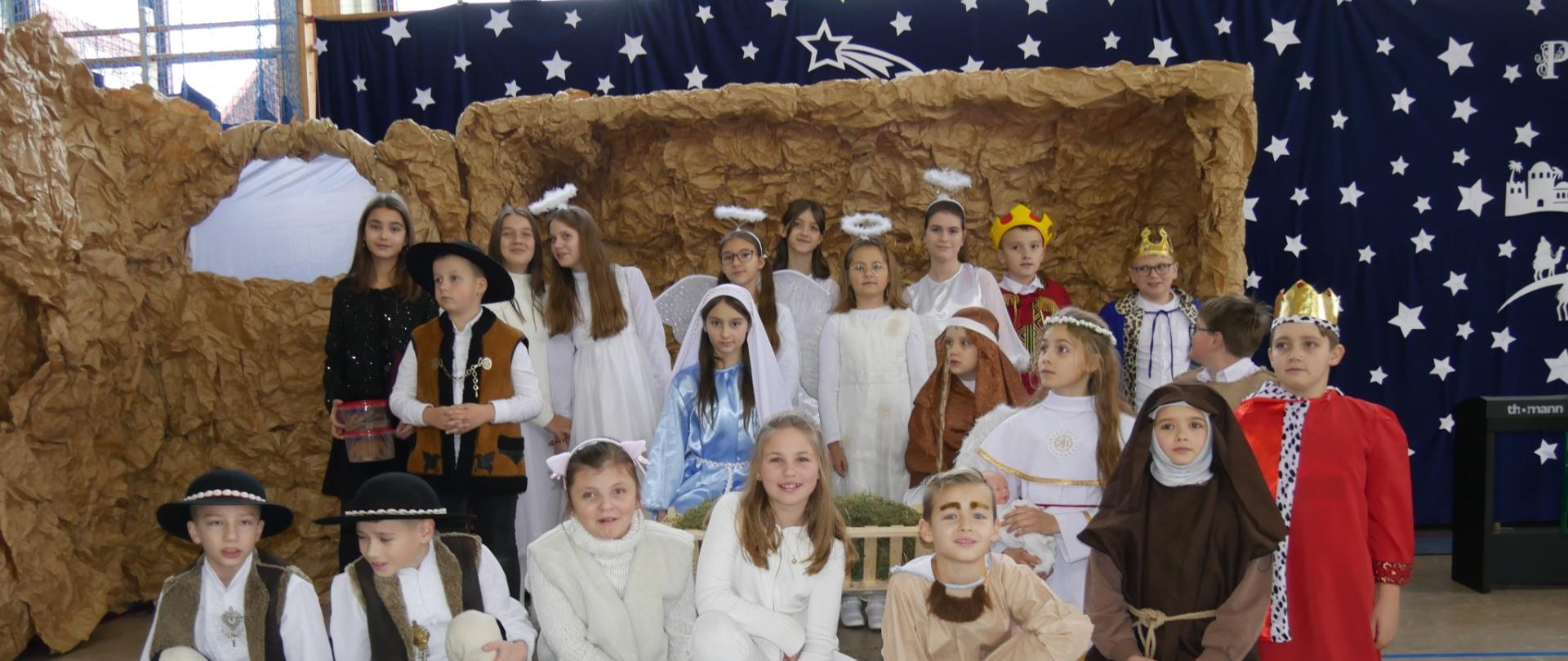 Uczniowie na tle szoki i dekoracji jasełkowej w strojach Matki Boskiej, Józefa, Trzech królów, aniołów, pastuszków.