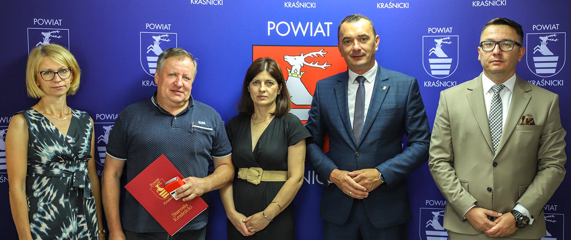 Pamiątkowe zdjęcie wykonane po podpisaniu umowy. Zdjęcie przedstawia pięć osób stojących obok siebie na tle niebieskiej ścianki reklamowej z wizerunkami herbu powiatu kraśnickiego.