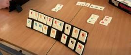 Bliskie ujęcie drewnianej ławki szkolnej, na której trwa rozgrywka w grę planszową Rummikub. Na pierwszym planie stoi czarna, dwupoziomowa podstawka wypełniona pionowymi, białymi kafelkami z kolorowymi cyframi (m.in. czerwone 1, czarne 3, niebieskie 4). Na środku stołu leżą ułożone przez graczy kombinacje kafelków w rzędach i grupach, a po prawej stronie widoczne jest czarno-czerwone pudełko z logo gry. W kadrze widać fragmenty rąk i postaci dwóch grających dzieci.
