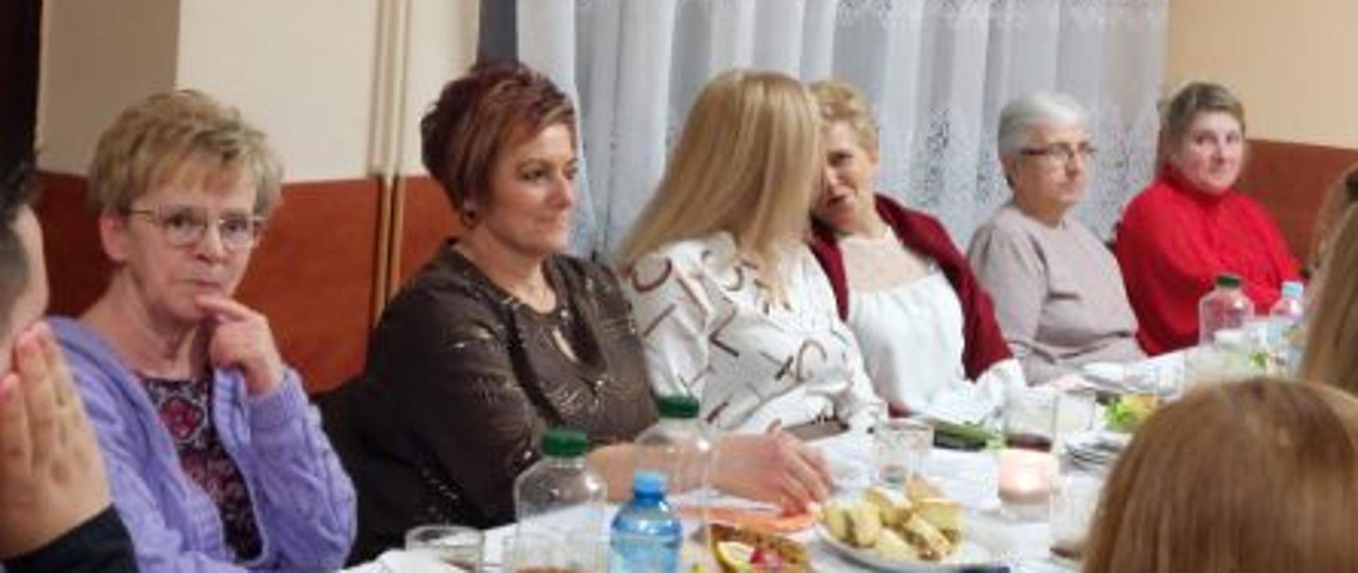 6 kobiet siedzących przy stole z białym obrusem. Na stole stoją napoje i jedzenie.
