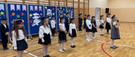 Uczniowie klasy 1 odświętnie ubrani przedstawiają program artystyczny na sali gimnastycznej, za nimi granatowa ścianka dekoracyjna z przypiętymi na nią papierowymi sylwetami drzewa, sowy, dziewczynki siedzącej na ołówku i kolorowymi liścmi