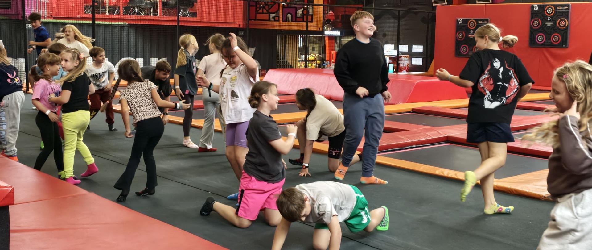 Dzieci podczas zabawy w Parku Trampolin Hangar 646 w Warszawie. Uczestnicy korzystają z trampolin, skaczą, wykonują różne ćwiczenia ruchowe i wspólnie spędzają czas w aktywny sposób. W tle widoczne są kolorowe strefy rekreacyjne oraz wyposażenie parku.