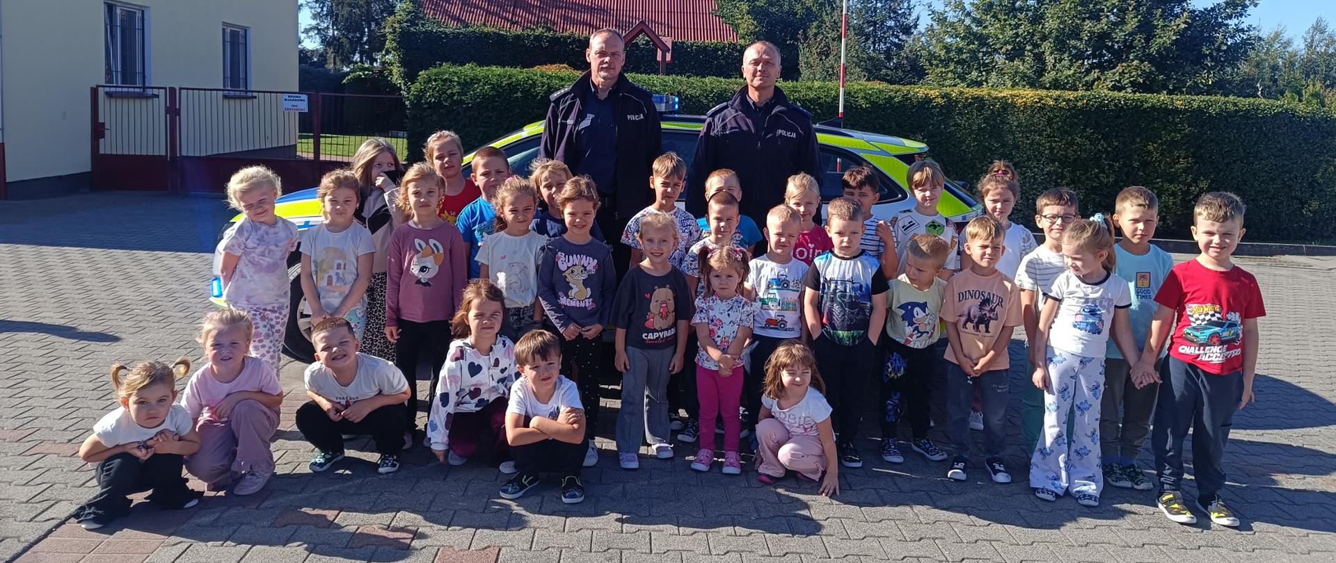 Zdjęcie przedstawia grupę przedszkolaków pozującą do zdjęcia razem z dwoma policjantami, za nimi stoi radiowóz. W tle widać drzewa, budynki, bramę i zielony żywopłot. 