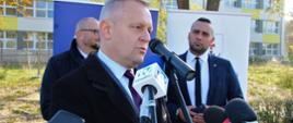 Mężczyzna stoi i udziela wywiadu do mikrofonów trzymanych przez dziennikarzy, w tle dwóch mężczyzn. 