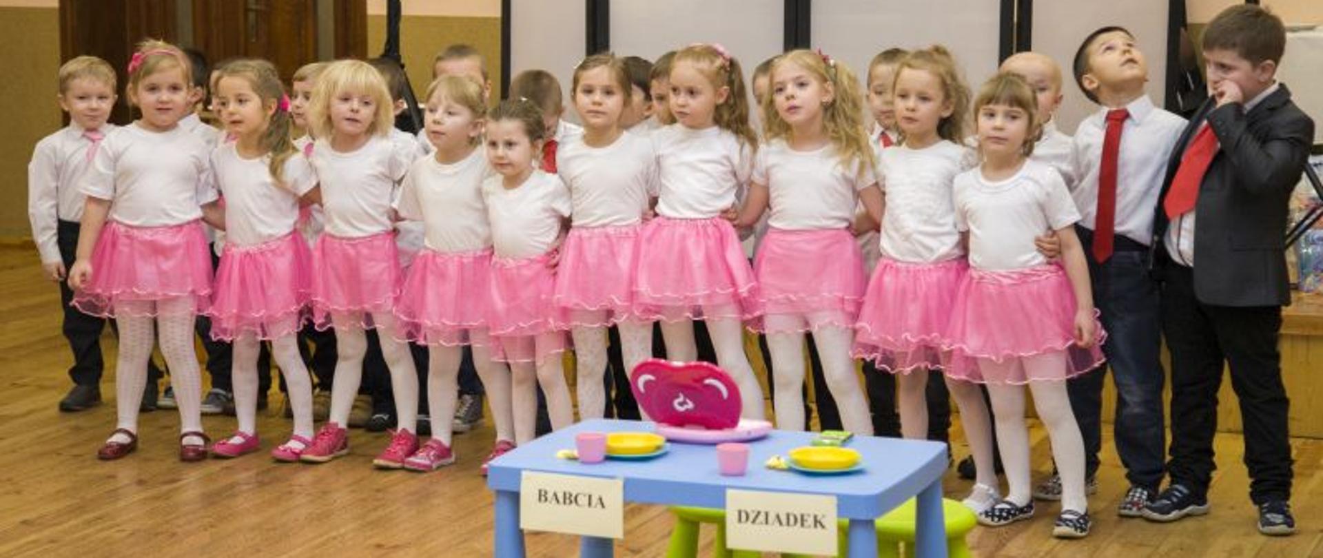 Grupa dzieci stojąca w dwóch rzędach, dziewczynki w różowych spódniczkach i białych bluzeczkach, chłopcy czarne spodnie, białe koszule, czerwone krawaty. Przed nimi niebieski stolik z przyklejonymi kartkami a na nich napis babcia, dziadek. 