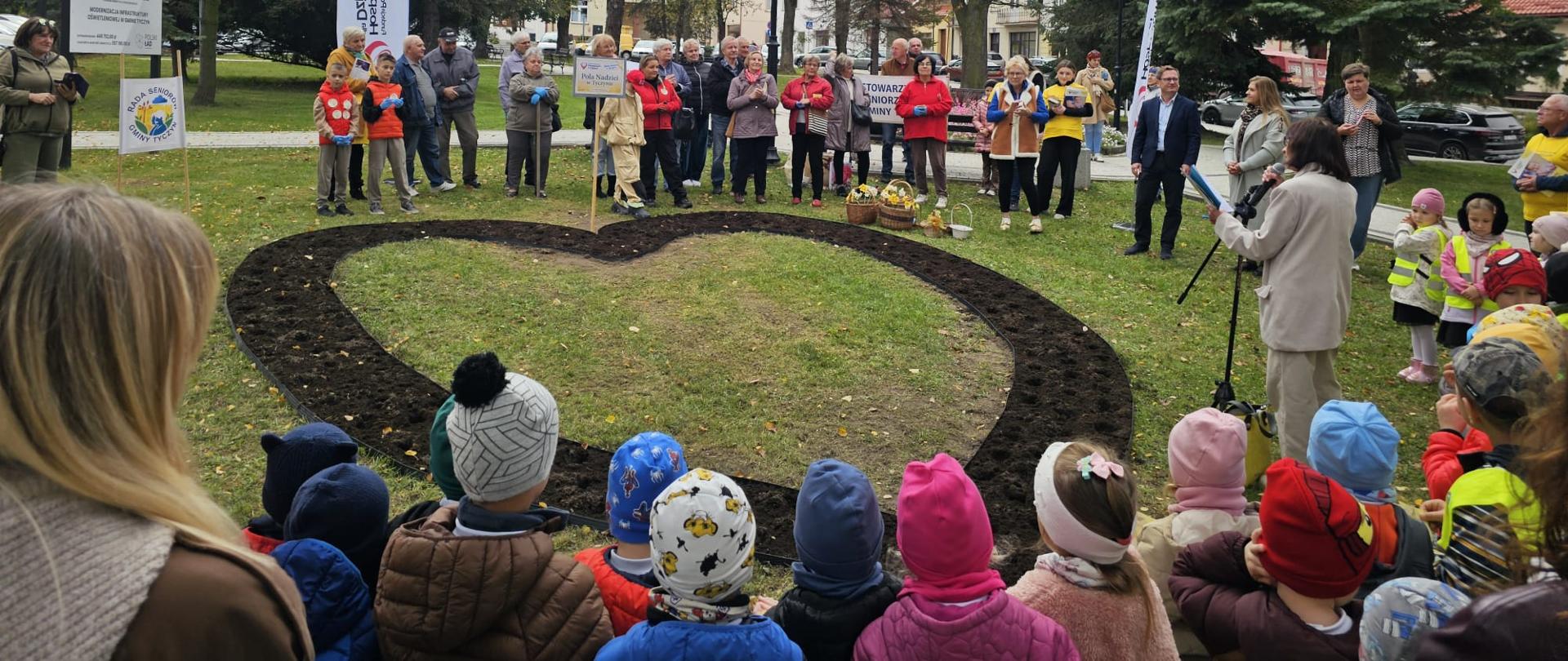 Na plantach w Tyczynie zgromadziły się dzieci i dorośli wokół rabaty w kształcie serca na której będą sadzone żonkile.