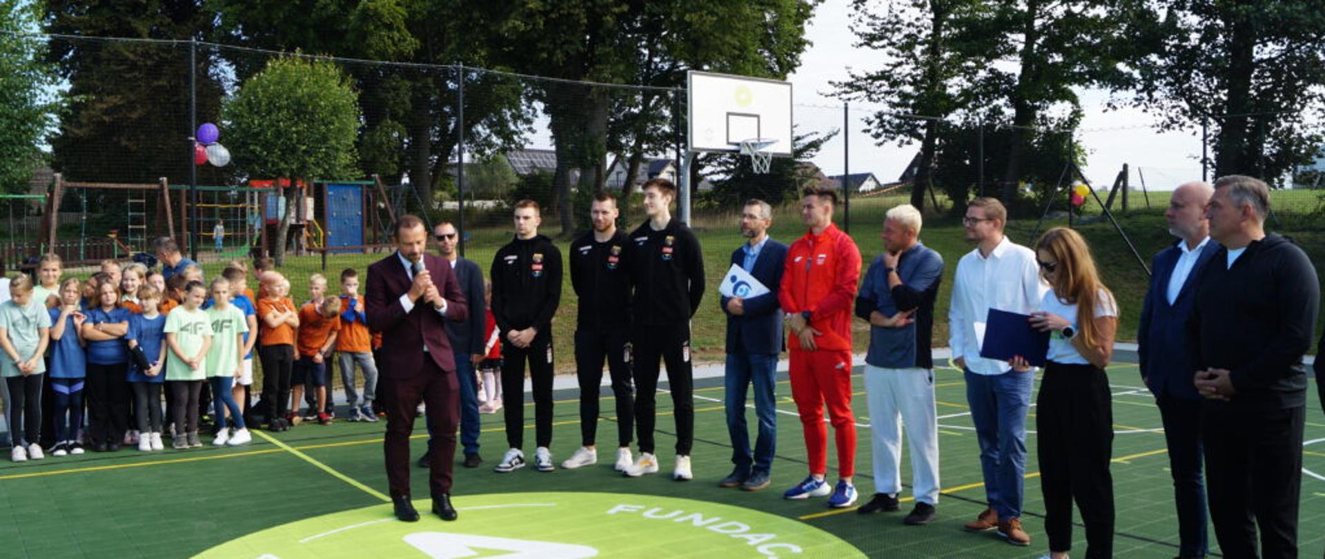 na zdjęciu znajduje się grupa ludzi i dzieci ubranych na sportowo, przemawia mężczyzna ubrany elegancko, wszyscy stoją na nowym, zielonym boisku
