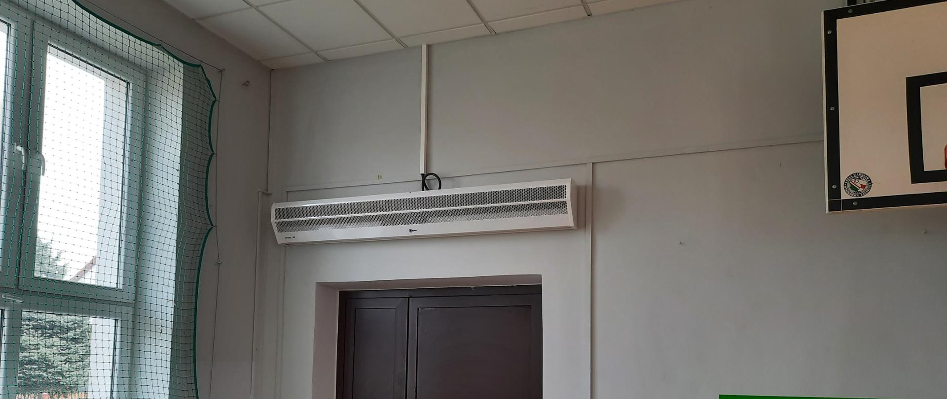 Sala OSP w Turzynie. Nad brązowymi drzwiami zainstalowana kurtyna powietrzna. Po lewej stronie okno zabezpieczone siatką. Pod oknem zielone krzesła.