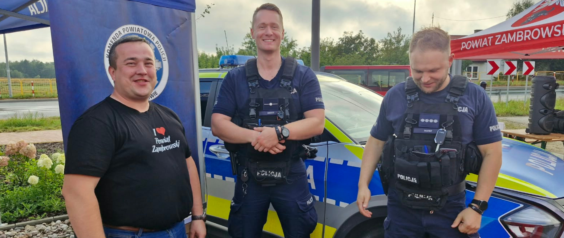 Radny powiatu zambrowskiego w koszulce promującej powiat stojący obok dwóch policjantów w pełnym oporządzeniu służbowym. W tle widoczny jest oznakowany radiowóz policyjny oraz namioty promocyjne.