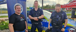 Radny powiatu zambrowskiego w koszulce promującej powiat stojący obok dwóch policjantów w pełnym oporządzeniu służbowym. W tle widoczny jest oznakowany radiowóz policyjny oraz namioty promocyjne.