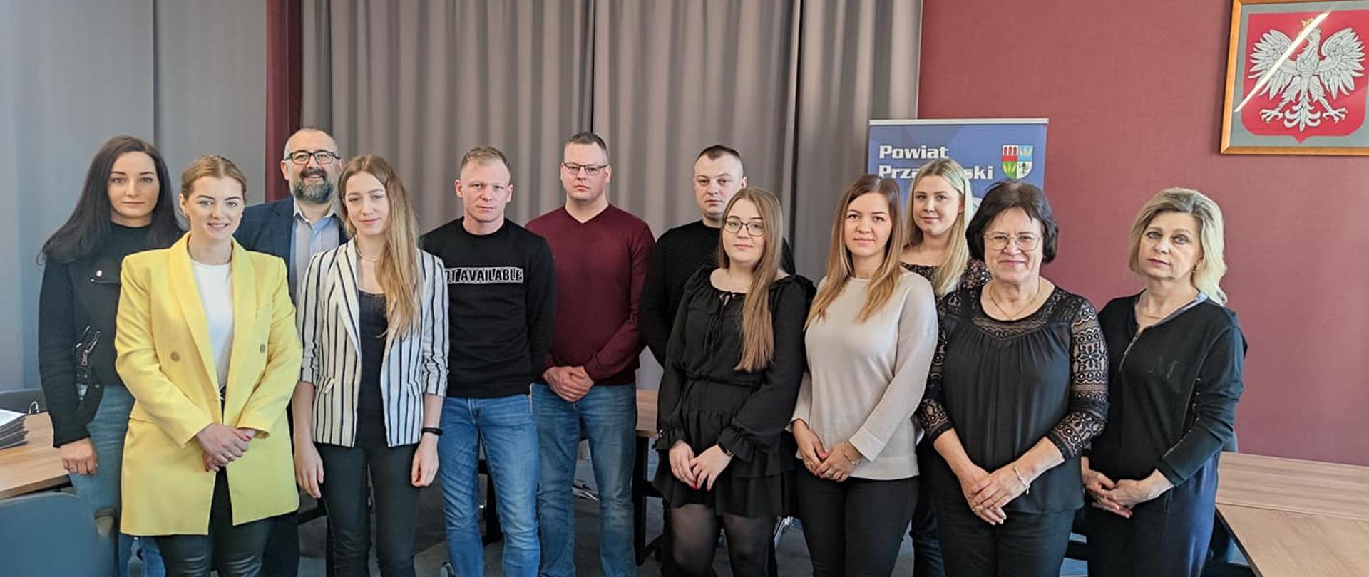 Zdjęcie przedstawia Starostę Przasnyskiego Krzysztofa Bieńkowskiego, Dyrektor Powiatowego Urzędu Pracy Jadwigę Żuber oraz Panią Barbarę Garlińską wraz z nowymi przedsiębiorcami powiatu przasnyskiego tuż po podpisaniu umów na dofinansowanie rozpoczęcia ich działalności gospodarczej. Zdjęcie wykonano na sali konferencyjnej Centrum Aktywizacji Przedsiębiorczości w Starostwie Powiatowym w Przasnyszu, w tle szare kotary, ściany w kolorze szlachetnego mahoniu oraz oprawione godło Rzeczypospolitej Polskiej – biały orzeł w złotej koronie na czerwonym tle.
