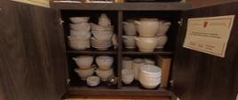Otwarte, ciemnobrązowe drewniane szafki w pomieszczeniu o drewnianych ścianach i podłodze. Wewnątrz uporządkowane są ceramiczne naczynia – talerze, miski i kubki – rozmieszczone na trzech półkach. Na górze znajdują się talerze i mniejsze miski, na środkowej półce większe naczynia, a na dolnej drobniejsze kubki i miski.