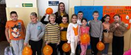 Dziesięcioro uczniów klasy trzeciej w pomarańczowych ubraniach stoi przy ścianie korytarza, na której zawieszone są plakaty z hasłami przeciwdziałania przemocy, uczniowie trzymają w rękach pomarańczowe balony, za dziećmi stoi wychowawczyni klasy