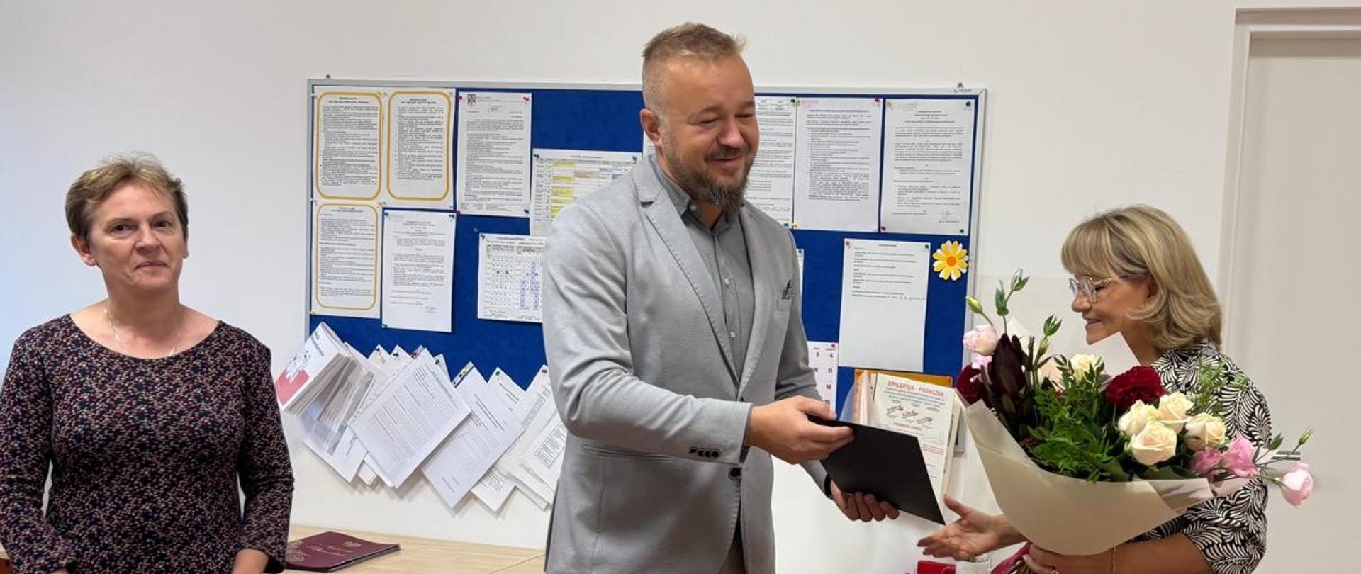 Wójt Gminy wręcza kwiaty i podziękowania Dyrektor Gminnego Przedszkola w Kobiórze z okazji Dnia Edukacji Narodowej. Obok stoi pracownik Urzędu Gminy.
