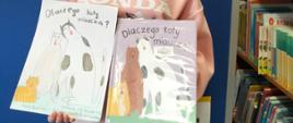 Dziewczynka z klasy 5 trzyma w jednej ręce wypożyczoną z biblioteki książkę przedstawiającą 3 koty, w drugiej ręce wykonany przez siebie obrazek z takim samym motywem