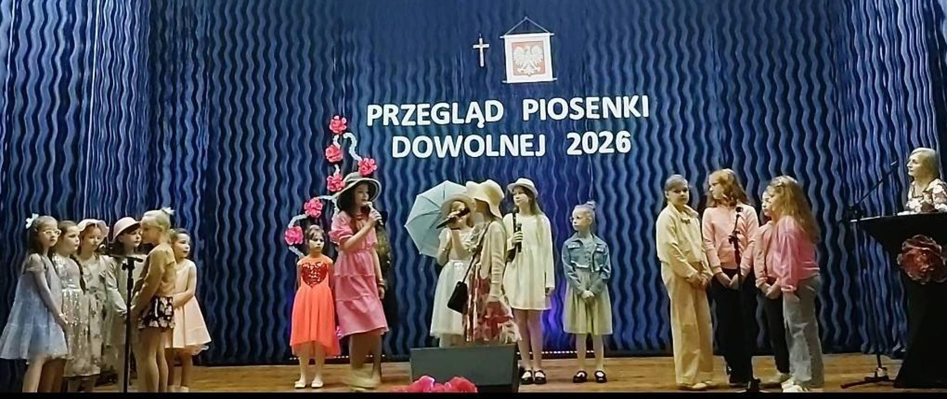 Na scenie znajduje się kilkanaścioro uczennic z klas młodszych. Uczennice ubrane są w kolorowe sukienki. Niektóre z nich mają na głowie kapelusze, a jedna z dziewczynek - parasol. Za uczennicami na granatowym tle wisi napis: PRZEGLĄD PIOSENKI DOWOLNEJ.