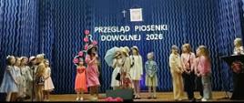 Na scenie znajduje się kilkanaścioro uczennic z klas młodszych. Uczennice ubrane są w kolorowe sukienki. Niektóre z nich mają na głowie kapelusze, a jedna z dziewczynek - parasol. Za uczennicami na granatowym tle wisi napis: PRZEGLĄD PIOSENKI DOWOLNEJ.